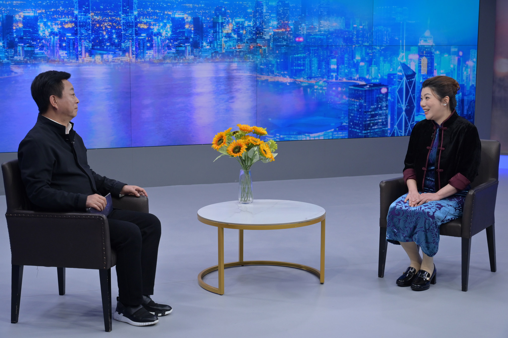 Two participants seated across a table during a studio conversation, with flowers and a harbour backdrop.