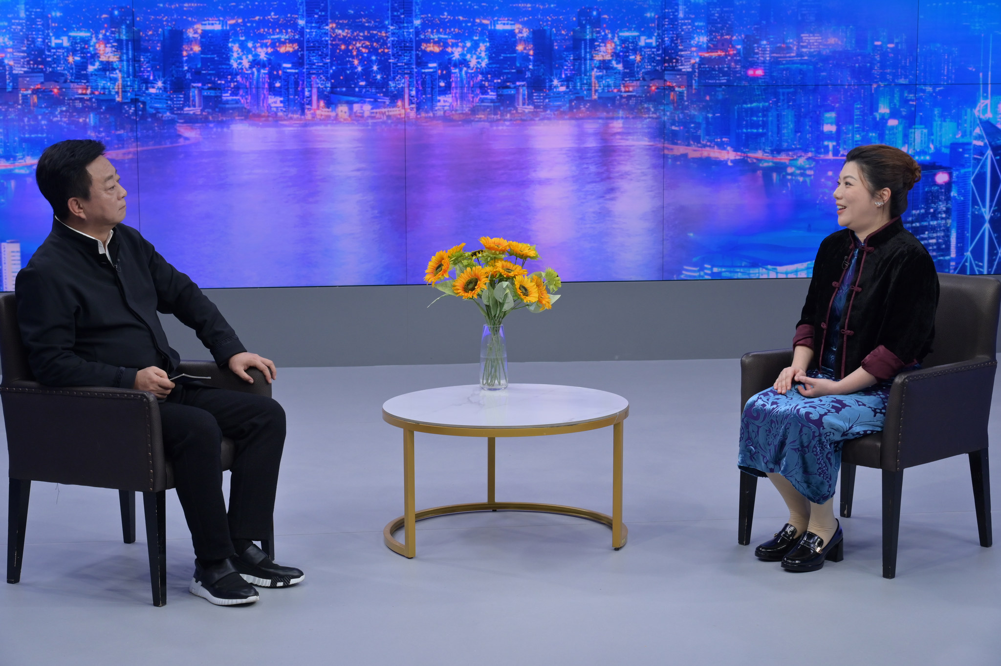 A second view of two participants in conversation in a studio, with a table, flowers and a harbour backdrop.